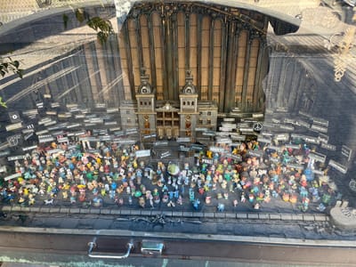 The image displays a window display featuring a miniature diorama. In the background is a detailed model of a grand building with multiple levels and ornate architectural elements, resembling a train station or a historical public building. The foreground is densely populated with numerous small figurines, predominantly cartoonish characters, animals, and whimsical creatures. These figurines are arranged in a crowded, layered fashion, creating a lively and somewhat chaotic scene. Many of the figurines are placed on a flat surface, while others are suspended by thin threads or wires, giving the impression of movement or a bustling crowd. Black signs with white text, some featuring symbols, are interspersed throughout the display, some attached to the figurines and others hanging independently. The overall composition suggests a whimsical, possibly satirical, or commemorative exhibit. The display is viewed through glass, which reflects the surrounding environment, including sunlight casting shadows and distorted images of the environment. The scene is framed by a dark, textured border at the bottom, which includes metal hardware resembling a suitcase latch and handle, suggesting the display might be within a suitcase or a similar container.
The Oulu 10 travel museum, located in Oulu, Finland, features this particular window display. The museum's exterior often showcases eclectic and artful window displays to attract visitors. This display, with its miniature figurines and signs, is a characteristic example of the museum's creative approach to exhibiting local history and culture.
