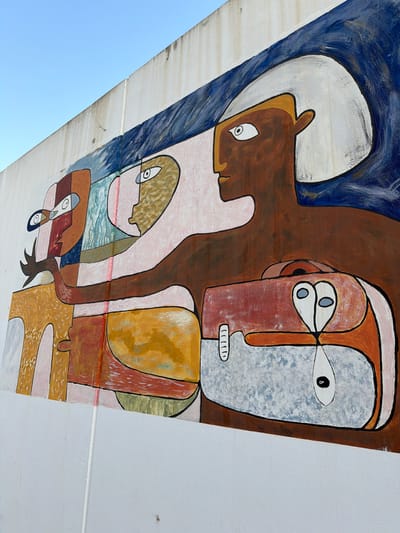 This is a close-up, low-angle shot of a colorful mural painted on a sloped surface, possibly a bridge or overpass, against a clear blue sky. The artwork features abstract figures with stylized faces and bodies, rendered in bold outlines and vibrant colors like brown, red, yellow, and blue. The style suggests a modernist or cubist influence.
In the upper left, a brown, horse-like head with large eyes is depicted within a swirling blue and white background. To its right, a large figure with a reddish-brown face and a textured light blue area for its eye dominates the composition. This figure has small, dark eyes and a mouth with white, linear teeth. Other figures and shapes, rendered in pink, yellow, and blue, are layered and intertwined, creating a sense of depth and complexity. One figure appears to be holding or interacting with another, smaller figure that has wide, dark eyes.
The mural appears to be outdoors, indicated by the clear sky and the way the light falls on the painted surface. The condition of the painted surface suggests it is exposed to the elements, with some signs of wear and tear. The context of Benimàmet-beniferri, Spain, implies an urban or semi-urban setting. The overall mood of the artwork is abstract and somewhat enigmatic, with the stylized figures evoking a sense of narrative or symbolic meaning that is open to interpretation. There is no visible text in the image.