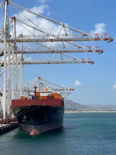 An anchored container ship is docked at a port in Ksar al Majaz, Morocco. The ship is a large cargo vessel with a red hull and black detailing, laden with numerous shipping containers stacked on its deck. Several large, white gantry cranes with red accents extend over the ship, indicating an active loading or unloading operation. The port facilities appear modern and extensive, with a pier made of dark wood or composite material. The water is a deep teal color, reflecting the clear blue sky dotted with white clouds. In the background, a hilly coastline with scattered buildings and vegetation suggests the proximity of the city. The overall scene depicts a busy commercial shipping hub under a bright, sunny sky.