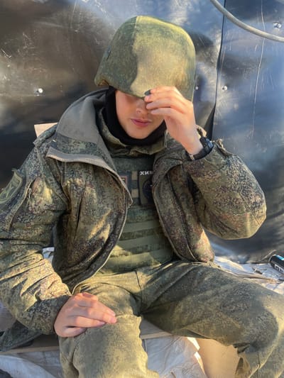 A person wearing a camouflage military uniform and helmet sits on what appears to be a wooden surface covered with white fabric. The person is looking down and has their right hand covering their eyes. The background consists of a dark, textured surface with some metallic elements and a rope. Patches are visible on the uniform, including one with Cyrillic text. The lighting suggests an outdoor setting with sunlight casting shadows. The person's left hand rests on their lap.