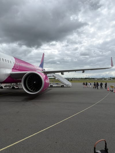 An overcast daytime scene at an airport tarmac in Tillé, France, shows a magenta and white Wizz Air aircraft. The image is taken from a low angle, looking upwards and along the side of the plane, which is rotated on its side.

The prominent subject is the large, single-aisle passenger jet, painted in Wizz Air's distinctive purple/magenta and white livery. The aircraft's right engine, with its visible fan blades, is in the foreground, and the fuselage extends towards the tail, featuring rows of windows. The right wing stretches into the distance, with a wingtip fence clearly visible. The text "WIZZAIR" is discernible on the side of the engine cowling, and "wizzair.com" can be vaguely made out on the wingtip.

To the right of the aircraft, several people are walking on the tarmac, appearing to be passengers or ground staff. They are heading towards or away from the aircraft, suggesting a boarding or disembarking process. A mobile boarding stair is positioned near the aircraft's entry door. In the far right foreground, part of a black stroller or pushchair handle is visible.

The ground is dark asphalt, with a light yellow line running diagonally across it, possibly indicating a safety boundary or taxiway edge. In the background, beyond the people, there are orange traffic cones and other airport equipment. The sky is heavily overcast with thick, grey clouds, indicating gloomy weather conditions.