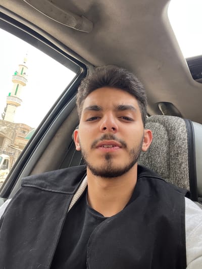 The image depicts a young adult male, likely in his 20s, with dark hair, a trimmed beard, and mustache, taking a selfie while seated in a vehicle. He is wearing a black open vest over a dark t-shirt and a light grey long-sleeved garment. The vehicle's interior shows a grey fabric headliner, a sun visor, and a portion of a grey textured seat cover. Outside the vehicle's left window, an urban street scene in Harasta, Syria, is visible under an overcast sky. A prominent white minaret, featuring three distinct levels with green-railed balconies and a green-capped spire, stands in the mid-ground. Behind the minaret are several light-colored buildings with flat roofs, and the front part of a white utility vehicle or truck is visible at street level. The male subject is looking directly into the camera.