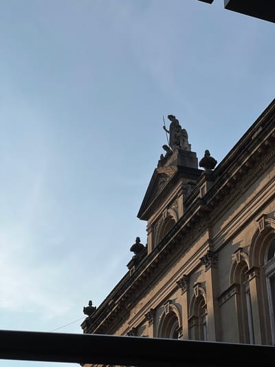 The image captures a close-up, low-angle shot of the facade of an ornate building, likely a historic public building, under a pale blue sky. The building's architecture features classical elements such as arched windows, decorative cornices, and sculptural embellishments. Atop the building, a statue of a figure holding a spear stands against the sky. The sunlight casts long shadows, indicating either early morning or late afternoon. The overall scene suggests an urban setting in Derry/Londonderry, United Kingdom, with the building's grandeur hinting at its significance within the city.
