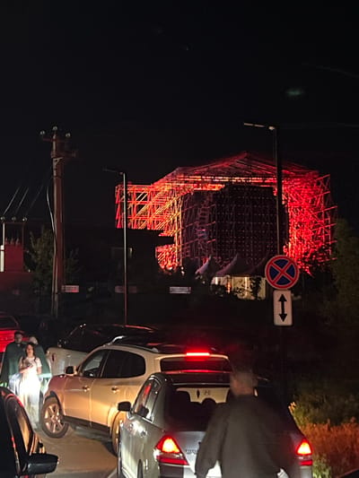 The image, taken at night in Jrvej, Armenia, shows an illuminated structure, appearing to be scaffolding, glowing red, possibly due to lights. A street sign prohibiting parking and allowing traffic in both directions is visible near the structure. Cars are lined up, indicating a possible traffic jam or obstruction. A person is present among the cars near a streetlight, but the image doesn't provide enough detail to determine the specifics of their activities or interactions. The environment is predominantly dark, with sparse lighting illuminating the immediate surroundings.