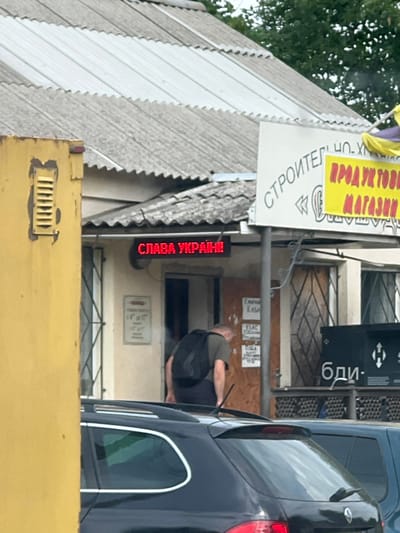 The image depicts a street scene in Martove, Ukraine. In the mid-ground, a single male individual is visible, wearing a dark green short-sleeved shirt and dark trousers, with a black backpack. He is bent forward, facing away from the viewer, appearing to lean into or over an object in the lower frame.

Behind the individual, a single-story building with a light-colored facade and a weathered corrugated metal roof is prominent. Above an open doorway on the left side of the building, a red LED display sign reads "СЛАВА УКРАЇНІ!" (Glory to Ukraine!). To the right of this entrance, another doorway appears to be boarded up with a wooden panel, onto which several handwritten signs are affixed, one partially legible as "КАСА" (Cashier). Further to the right, a window with a diamond-patterned security grille is visible. Above the building, a white sign with black text partially reads "СТРОИТЕЛ" (Builder), and a yellow sign below it displays "ПРОДУКТОВИЙ МАГАЗИН" (Grocery Store).

In the foreground, the rear section of a dark-colored SUV or wagon with a roof rack and rear wiper is visible, partially obscuring the individual. To the right of this vehicle, the front portion of a light blue vehicle is also present. On the far left of the image, a large, yellow, rectangular structure with a horizontal vent is visible. Green tree foliage and a cloudy sky are discernible in the upper background.