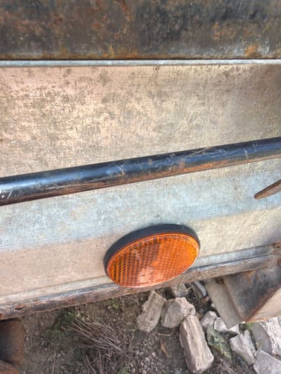 A close-up, slightly low-angle shot shows the side of a weathered trailer in Ust'-Dzhigutinka, Russia. The trailer's exterior is a dull, galvanized metal with visible scratches and rust. A black metal pipe, also showing signs of rust, runs horizontally across the frame. Below the pipe, an orange, oval reflector with a black bezel is attached to the trailer. The reflector has a textured surface with a repeating pattern and some faint white text is visible on its surface. The ground beneath the trailer is composed of loose gravel, dirt, and some dried plant debris, with a few scattered rocks. The lighting suggests it is daytime. No people or animals are visible in the frame.