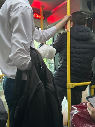 The image depicts the interior of a public transportation vehicle, likely a bus, with passengers. A person on the left wears a white collared shirt and dark trousers, with a black backpack hanging from their left shoulder. Their right arm is bandaged. To the right of this individual, another person is visible from behind, wearing a dark puffer jacket and holding onto a yellow handrail. The bus interior features red illuminated signage above the handrails, and a window shows a glimpse of greenery outside. A person on the far right is holding a mobile phone. The scene suggests an ordinary commute. No specific indicators confirm the location as Smolenshchina, Russia.