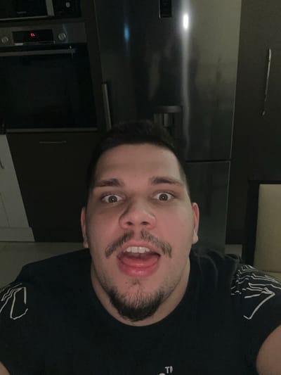 The image depicts a single adult male in an indoor setting. The specific location in the city of Radishevo, Bulgaria, is not discernible from the visual evidence provided. The man, positioned centrally in a close-up, is looking directly at the camera with wide-open eyes and an open mouth, his tongue slightly extended. He has short dark hair and facial hair consisting of a mustache connected to a goatee. He is wearing a black t-shirt featuring white graphic patterns on the shoulders. In the background to the left, dark kitchen cabinetry is visible, along with an oven or microwave unit displaying "CL" on its digital interface and control knobs. To the right, a stainless steel refrigerator with a water/ice dispenser is present, reflecting ambient light. A dark wall with a vertical light-colored mark and a portion of light-colored furniture are visible in the far right of the frame. The scene is moderately lit.