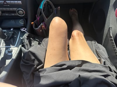 The image, likely taken from an elevated viewpoint inside a vehicle, shows the interior of a car. In the driver's seat, the legs of a person wearing a black garment are visible. The left leg appears to be a prosthetic, positioned prominently in the foreground. A brown leather handbag with blue and purple accents is near the gear shift. The dashboard, steering wheel, and center console features are visible, as well as what appears to be a pair of sunglasses in a case. The location is unidentifiable from the available visual information; nothing in the image suggests that the scene is located in Leidschendam, Netherlands.