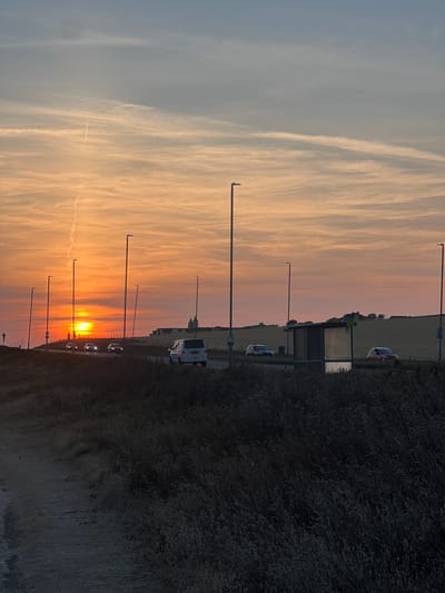 The image depicts a road scene at sunset in Ovingdean, United Kingdom. A bright orange sun is low on the horizon, casting a warm glow across the sky, which transitions from orange to pink and then to a pale blue. Wispy clouds, some with contrails, are scattered across the sky.
Several cars are visible on the road, including a white van on the right side, and three cars in the distance driving towards the sunset. There are multiple tall lampposts lining the road. On the right side of the road, a bus shelter with glass panels stands beside the road. In the background, rolling hills and buildings can be seen, suggesting a rural or semi-rural setting. The foreground is dominated by dark, silhouetted vegetation, possibly bushes and long grass, along a dirt path on the left. The overall impression is a peaceful, end-of-day scene on a road leading towards a picturesque landscape.