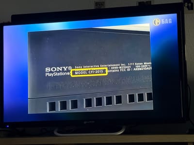 This is a photograph of a television screen displaying information about a Sony PlayStation. The screen shows the "SONY" and "PlayStation®" logos, along with the model number "MODEL CFI-2015". Text below indicates it contains FCC ID: AK8M21DAU1 and is rated for 100-240V~. A logo with the letters "G БАГ" in a circular emblem is visible in the upper right corner of the screen. The television is placed on a wooden surface, and some cables are visible below it. The background suggests an indoor setting. There is no information in the image that can be used to determine the location of Castillo Bajo-conejito, Spain.