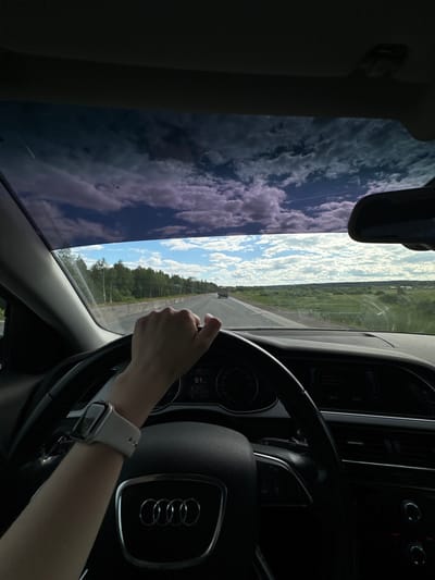 This media file depicts a first-person perspective from inside a car, likely an Audi given the prominent emblem on the steering wheel. The scene captures a drive through a rural landscape in Yus'ki, Russia. The image is oriented vertically, suggesting the photo was taken while the car was in motion.
The primary subject is the driver's hand, resting on the steering wheel, and their forearm, adorned with a smartwatch. Through the windshield and side window, a bright, cloudy sky is visible, reflecting on the tinted glass. The landscape outside consists of lush green fields and trees, with a road stretching into the distance. A faint contrail from an airplane can be seen in the sky.
The dashboard of the car is partially visible, showing a digital display with the speed indicated as 61. Other text on the display is too small to be legible. The time displayed is 10:03. The weather appears to be pleasant, with a mix of sun and clouds, suggesting a daytime activity. The overall mood conveyed is one of serene travel, perhaps a leisurely drive through the countryside. The setting appears to be a paved road, possibly a highway or a main route, indicating travel between locations.