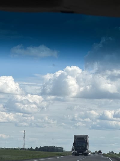 The image depicts a road scene from a vehicle's perspective, oriented vertically. A dark, possibly tinted, window frame is visible on the left side. The sky is a dominant feature, filled with a mix of bright blue sky and dramatic, billowing white clouds.
In the distance, a highway stretches out, with several vehicles visible. Prominently, a dark-colored truck is driving on the right side of the road. Further ahead, other vehicles, including a lighter-colored truck and a white car with illuminated headlights, are also traveling. The road appears to be a multi-lane highway, indicated by the white lines marking the lanes.
To the right of the road, a green grassy field borders a line of trees. In the middle ground, a tall, skeletal tower, possibly a cell tower or radio tower, stands against the cloudy sky. The overall scene suggests a daytime drive, likely during pleasant weather, with the sun filtering through the clouds. The location is identified as Акцябрскі сельскі Савет, Belarus. No text is visible in the image.