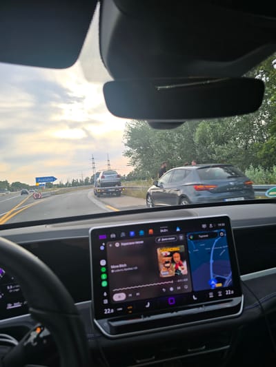 The image depicts a view from inside a car driving on a highway in Bischofsheim, Germany. The car's dashboard displays a touchscreen infotainment system with navigation and music playback. Outside, a tow truck is seen on the right shoulder of the highway, carrying a damaged car. The highway is bordered by trees and utility poles. The sky is cloudy with the sun setting, casting a warm glow. Several other vehicles are visible on the highway in the distance. The sign on the left indicates "Ausfahrt" (exit) with an arrow pointing to the right. The scene appears to be captured during daylight, likely in the late afternoon or early evening.