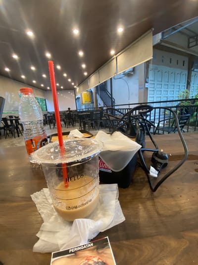 This image, set in Keudeublang, Indonesia, shows a close-up of a wooden table in what appears to be a cafe or co-working space. The primary focus is on several items resting on the table.
In the foreground, a plastic cup containing a light brown beverage, likely coffee, is visible. The cup has "HELLO" printed on it and an Instagram handle "@68.coffeeshop". A red straw protrudes from the cup, which is surrounded by crinkled white tissue paper. Next to the cup is a clear plastic bottle of water with an orange cap and a partially visible label that includes "Claro".
Further back on the table, a black power adapter or charger is visible with tangled cables. A piece of paper or magazine page with the word "PERINGATAN" (meaning "WARNING" in Indonesian/Malay) is also present.
The background reveals more of the interior space. There are numerous small, bright lights embedded in the ceiling, illuminating the area. Beyond the table, an array of dark chairs and tables suggests a seating arrangement. Through a window or open area in the distance, parts of an exterior building with white, paneled doors or windows are visible, along with some greenery and a yellow industrial tank. The scene suggests a relaxed atmosphere, possibly a break or work session at a cafe.