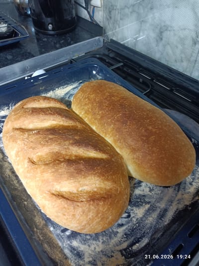 The image shows two loaves of freshly baked bread on a dark blue baking tray. The bread is golden brown with visible scoring marks on the crust of the loaf in the foreground. The background shows a kitchen counter with a black appliance, possibly a kettle, and a tiled wall. The scene is lit from above, casting shadows on the bread. The timestamp "21.06.2026 19:24" is visible in the bottom right corner of the image. There is no information in the image to suggest a location in Chuy, Kyrgyzstan.