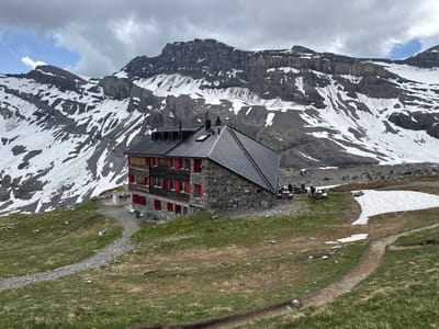 The image shows a stone and wood mountain hut with red shutters, set against a backdrop of snow-covered mountains and a cloudy sky. The hut is located in a grassy, sloped terrain with a gravel path leading towards it. In front of the hut, there are picnic tables with people seated, and a pile of laundry hanging on a line. Further in the background, there are stacks of stones and some patches of snow on the mountainside. The environment suggests a remote, high-altitude location, consistent with the area around Leukerbad, Switzerland, known for its alpine scenery and mountain huts.