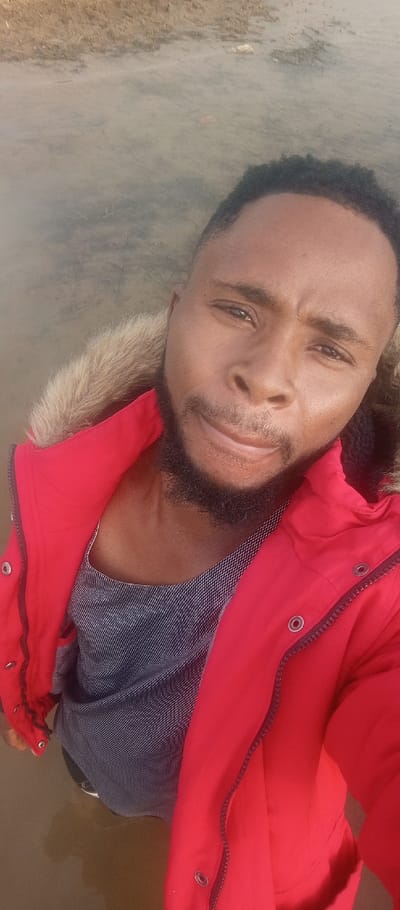 A man stands in shallow, murky water, taking a selfie from a low angle. He has dark skin, a beard, and short, dark hair. He's wearing a gray tank top and a bright red jacket with a fur-lined hood. The background is blurred but appears to be a muddy shoreline.
