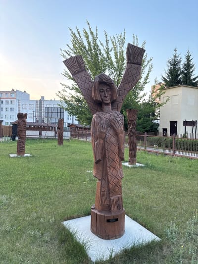 In Olchowa Kładka, Poland, an outdoor scene features several wooden sculptures in a grassy area. The primary sculpture in the foreground is a tall, carved wooden figure with a patterned robe and outstretched, wing-like appendages. In the background, there are apartment buildings, a fence, trees, and smaller wooden sculptures. The sky is clear and light blue, suggesting it is daytime, possibly late afternoon or early evening due to the soft lighting. The ground is covered in green grass with some patches of dandelions and other small white flowers. A dark plaque is attached to the base of the main sculpture.