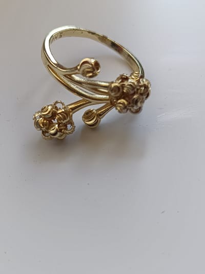 A close-up, high-angle shot shows a gold-toned ring with a floral or bud-like design. The ring is an open-ended bypass style, with two decorative elements, each resembling a cluster of small, curled petals, extending from opposite ends of the band and overlapping. The surface appears to be a smooth, light-colored material, possibly a table or countertop. There is no indication of any location within Arimjan, Kyrgyzstan, or any people, actions, or interactions. The image focuses solely on the object and its immediate surroundings.