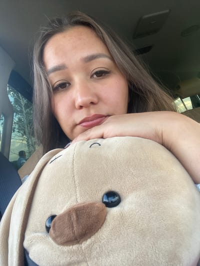 In Staromusino, Russia, a woman is taking a selfie in a car. She is holding a tan plush toy dog. The woman is looking at the camera with her head resting on the toy. The interior of the car is visible behind her, including the headliner and a window showing trees and another person in the distance.