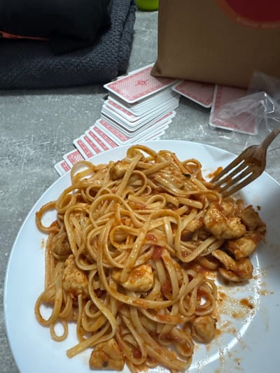 A high-angle, close-up photograph depicts a meal and miscellaneous items on a grey, textured surface, likely indoors in Krems an der Donau, Austria. A white, circular plate, centrally positioned, holds a serving of linguine pasta with diced chicken in a red-orange tomato sauce; a light-colored fork is inserted into the pasta on the right side of the plate, coated with sauce. In the background, to the left, a dark grey folded towel or blanket rests on the surface. To the right of the towel, multiple stacks of playing cards with red backs, each featuring a circular design, are fanned out and piled on the grey surface. Behind the cards, a light brown paper bag with a partially visible red circular logo stands upright. A crumpled clear plastic bag is positioned to the right of the paper bag. A small, bright green object is faintly visible in the upper left background. No people are present in the scene.