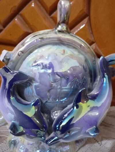 A glossy ceramic figurine features a globe encased within a round, lidded container. Several stylized dolphins, colored with iridescent blue and hints of yellow, adorn the base and appear to support the globe. The lid has a knob on top, and the entire piece has a shiny, reflective surface. A small chip is visible on the left side of the lid. The background shows a textured surface.