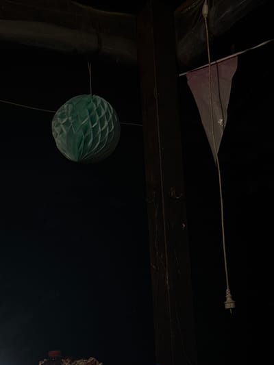 This image captures an outdoor setting at night in Gradets, Bulgaria. The primary subjects are decorative elements hanging from what appears to be a covered outdoor space, possibly a porch or gazebo.

In the upper portion of the image, a light-colored, kite-like object made of plastic sheeting is suspended by a thin wire. The wire extends to a white electrical plug, suggesting it's part of a light string. A dark, horizontal beam runs across the middle of the frame, providing a structural element. Below this beam, a round, teal-colored honeycomb lantern hangs by a string. To the left of the lantern, a dark, cylindrical object, possibly a pole or column, is partially visible. The background is predominantly black, indicating nighttime.

There is no activity or event clearly depicted; the scene is static, focused on the suspended decorations. Notable details include the nighttime setting and the artificial lighting implied by the electrical plug. The overall mood is subdued and possibly festive due to the presence of decorations, though the darkness and isolation of the elements create a somewhat stark impression. No visible text is present in the image.
