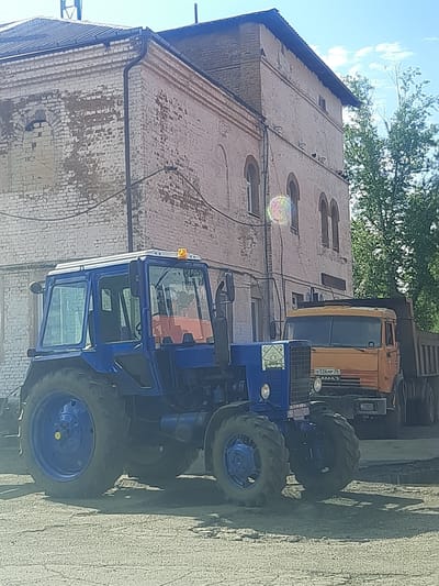 The image depicts a blue tractor and an orange dump truck parked on an asphalt surface in front of a brick building. The building appears to be an older structure with peeling paint and arched windows. The tractor is in the foreground, and the dump truck is partially visible behind it. Trees are present in the background. A license plate on the dump truck reads "PO 526 MP 38" and "KAMA3". The scene is set in the city of Shelekhov, Russia, on a bright, sunny day. There are no people visible in the image.