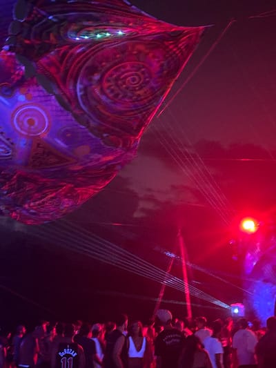 An outdoor night scene in Ban Mae Ping, Thailand, depicts a gathering of people illuminated by artificial lighting and laser effects. In the upper left quadrant, a large, multi-textured fabric banner with intricate spiral and geometric patterns, predominantly in purple and magenta hues, hangs suspended, with small green and white lights embedded along its upper edge. The background features a dark sky with faint cloud formations. Multiple white laser beams emanate from the right, traversing the frame diagonally downwards and to the left. A prominent, intense red spotlight is positioned on the mid-right, casting a strong red glow across the scene, with a supplementary blue light source visible below it. The lower half of the image is occupied by a dense crowd of people, largely silhouetted, standing and observing. Identifiable details among the crowd include a person wearing a dark jersey with "DEROZAN 11" on the back in the lower left, a person in a white top in the center, and an individual wearing a dark shirt with a "Patagonia" cap in the lower right. Support structures are faintly visible in the background, consistent with an event setup.