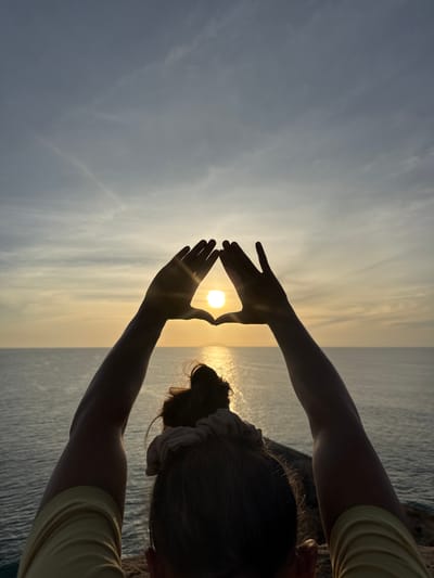 This is a portrait-oriented photograph taken at sunset in Shastrawela, Sri Lanka. The image features a person, silhouetted against the bright sun and the ocean. The person's head and arms are visible from behind, with their hair tied up with a scrunchie. They are holding their hands up to form a triangle, framing the setting sun. The sun itself is a bright, golden orb, casting a warm glow on the scene. The ocean stretches out to the horizon, with gentle waves visible. The sky is a soft blend of blues and yellows, with wispy clouds that indicate a clear but overcast sky. The overall mood of the image is peaceful and contemplative, with the person seemingly embracing the beauty of the sunset. There is no visible text in the image.