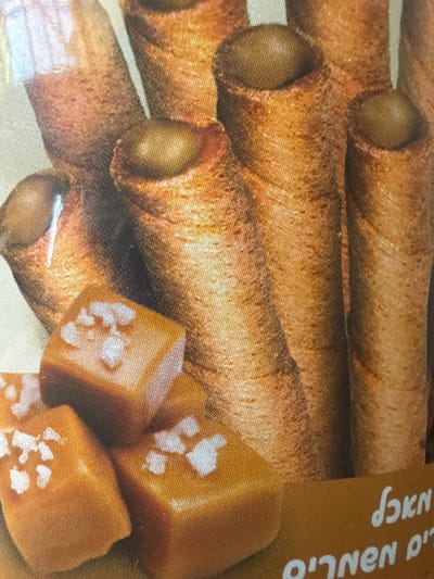 This image is a close-up, overhead shot of a pile of chocolate-filled wafer rolls and some caramel cubes sprinkled with sea salt. The wafer rolls are elongated and have a golden-brown, crispy texture, with visible chocolate filling at the ends of some. The caramel cubes are a rich, amber color and are scattered among the rolls.
The image also features some text in Hebrew on a light brown background, appearing on the right side of the frame. The visible words are "נחשונים" (Nakhshonim) and "קפה" (Cafe), suggesting a coffee-flavored product or brand.
The lighting is somewhat diffused, creating soft shadows and highlighting the textures of the food. There are no people or any indication of a specific time of day or weather. The overall impression is that of a product shot, possibly for packaging or advertising, emphasizing the sweet and indulgent nature of the treats. The location context of Dimona, Israel, does not provide any discernible visual cues within the image itself.