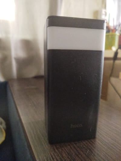 A black rectangular electronic device with a white stripe across the top sits on a wooden surface. The device has the "hoco" logo embossed on its lower portion. In the background, a sheer curtain is visible, and further back, out-of-focus objects suggest an indoor setting. The image does not provide any information about the city of Malooleksandrivka, Ukraine, or any people, actions, or interactions.