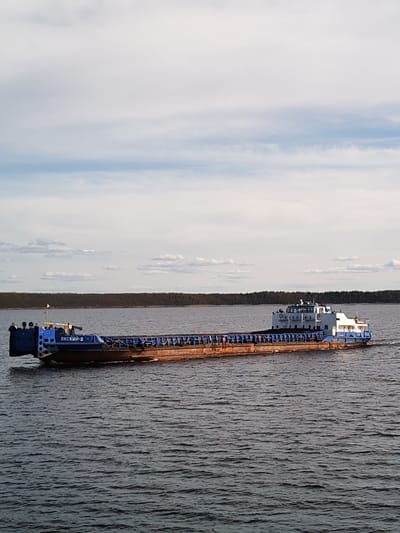 A barge labeled "OKCKNN-2" is sailing on a body of water in Gorushka, Russia. The barge is primarily rust-colored with blue sections at the front and rear. A cabin-like structure is visible at the rear. The water is choppy. A strip of trees lines the horizon in the background, and a cloudy sky occupies the upper half of the image.