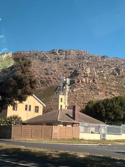 The image depicts a scene in Muizenberg, South Africa. A large, rugged mountain dominates the left side of the frame, its rocky terrain covered with sparse vegetation. Adjacent to the mountain, there is a telecommunications tower and a group of buildings. The buildings appear to be residential. A fence and a paved road run parallel to the buildings, bordering the right side of the image. The sky is a clear blue. The scene is brightly lit, suggesting it is daytime.