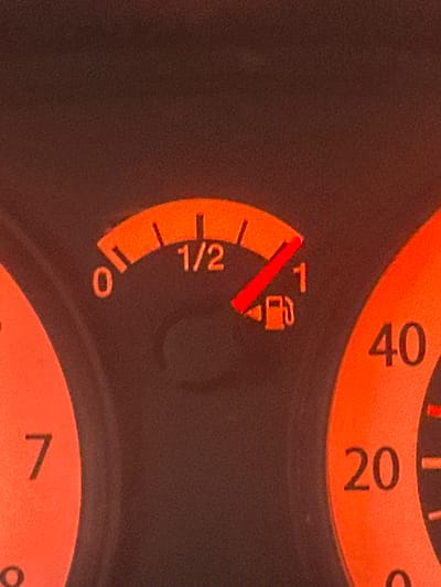 The image shows a close-up of a car's dashboard, specifically the fuel gauge and part of the speedometer. The fuel gauge is illuminated in orange and shows the needle pointing approximately three-quarters of the way between the '1/2' and '1' marks, indicating the fuel tank is more than half full. The markings on the fuel gauge range from '0' to '1'. To the left of the fuel gauge, a portion of another dial is visible with the number '7' at the top. To the right, a section of the speedometer is visible, with the numbers '40' and '20' clearly displayed. The background is a dark, textured surface, likely the dashboard material. The lighting suggests it is dark outside or the car's interior lights are on. There are no people visible in the image, and no information about the location in Tomilova, Russia is present in the visual data.