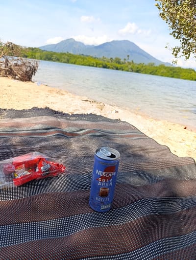 A can of Nescafe Gula Aren coffee sits on a striped blanket. The blanket is spread out on a sandy beach. The beach is located on the coast of the island of Java, Indonesia, near the village of Banyuputih, with coordinates -7.749 latitude and 114.298 longitude. In the background, a hill can be seen. A pile of wrapped candy lies on the blanket next to the can. A branch of a tree extends into the upper right corner of the picture, with its leaves out of focus. The sky is blue and clear. The water of the sea is calm and blue. The sand is beige and slightly damp. There is no sign of people in the picture.