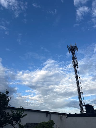 A cell tower extends diagonally across a blue sky filled with white, wispy clouds. The tower is made of a dark, lattice-like metal structure and is topped with a circular dish and several smaller antennas. The bottom right of the image shows the side of a white building with a dark, sloped roof. Green foliage is visible at the bottom right corner of the building. The image appears to have been taken outdoors on a clear day, possibly in the morning or late afternoon, given the bright sky and clouds. There is no text visible in the image. The scene depicts a telecommunications structure against a natural backdrop, suggesting an urban or semi-urban environment.