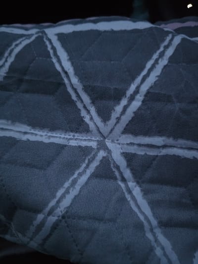 The image shows a textile pattern featuring a central starburst design with thick, light-colored lines radiating outward. These lines appear textured or slightly frayed, contrasting against the darker fabric base. The background showcases a quilted effect with subtly visible geometric shapes, possibly cubes or diamonds, adding depth to the overall design. The lighting suggests a soft, possibly artificial, illumination, highlighting the texture and pattern.