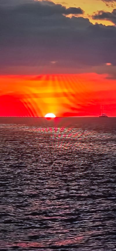 The image depicts a sunset over a body of water, likely the sea or a large lake. The sun, a bright orange orb, is partially submerged below the horizon, casting a vibrant red and orange glow across the sky. Rays of light fan out from the sun, creating a dramatic effect against the darker, purplish clouds above. On the right side of the frame, a small sailboat with two masts is visible on the water, silhouetted against the colorful sky. The water's surface is rippled, reflecting the colors of the sunset. There is no discernible location in Lippstadt, Germany visible in this image.