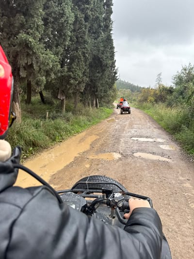 The image depicts a first-person perspective from an individual operating an all-terrain vehicle (ATV) on a muddy, unpaved road in Çiftlikköy, Turkey. The driver wears a red helmet, partially visible on the left, and a dark, possibly padded, jacket, with one hand gripping the right handlebar of the ATV. Ahead, two other ATVs are in a procession: the lead ATV is orange, followed by a maroon ATV. The rider of the maroon ATV is covered by a light-colored, possibly white, hooded garment. The dirt road is extensively marked with tire tracks and numerous large puddles of brown water, indicating recent moisture. To the left, a dense line of tall, slender, dark green coniferous trees borders the road, with an undergrowth of grass and smaller vegetation. The right side of the road features varied green and yellowish-brown foliage. In the distant background, a tree-covered hill is visible. The sky is uniformly overcast and light gray.