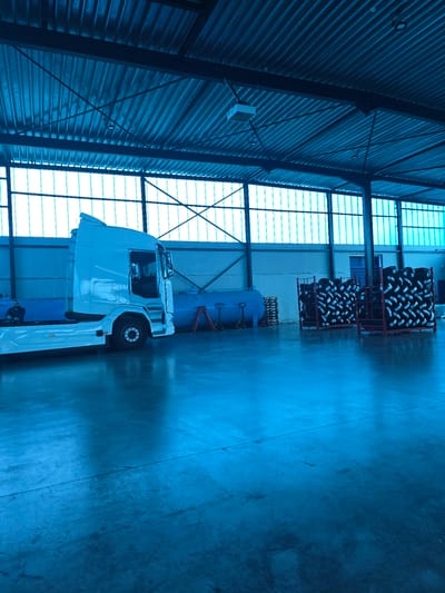This media file depicts the interior of a large industrial warehouse or garage, bathed in a strong blue light that gives the entire scene a monochromatic, cool tone.

The main subject is a white semi-truck (tractor unit), parked on the right side of the concrete floor. Behind the truck, a large, blue cylindrical tank, likely an air compressor or storage vessel, is visible. Further in the background, two red metal racks are stacked high with numerous new tires, indicating storage or inventory.

The building's structure is characterized by corrugated metal walls and ceiling on the left side, interspersed with translucent panels that allow ambient light to filter in. The floor is made of concrete, showing some wear and lines. A small white rectangular object is affixed to the corrugated wall on the left. A window or opening is also visible in the distant wall behind the tire racks.

The scene is static, portraying an industrial space used for vehicle storage, maintenance, or parts storage. No people are present, and no specific activity or event is actively taking place. The strong blue color cast is a notable detail, making it difficult to determine the exact time of day or external weather conditions. There are unreadable white labels on the tires, but no other discernible text is visible within the image. The overall impression is one of a vast, utilitarian industrial environment.