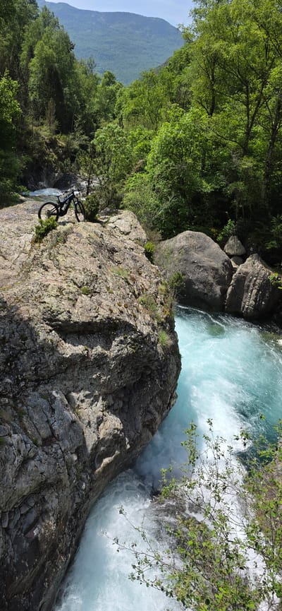 The image shows a mountain bike positioned on a rocky cliff edge, seemingly parked to overlook a rapid river. The cliff face is composed of layered gray and tan rock with patches of green moss. The river is a churning mix of turquoise and white water, suggesting a powerful current. Green foliage, including trees and shrubs, surrounds the rocky area. A mountain range is visible in the background. The location is Sahún, Spain.