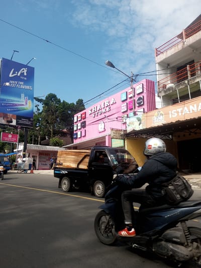 The image captures a street scene. A blue scooter with a rider wearing a helmet, a dark jacket, jeans, and red-and-white sneakers is in motion on the right side of the frame. A black truck with an open bed is directly behind the scooter. Behind the truck, a pink building with the words "KHAIRA STORE" and various brand logos is visible. To the left, a large billboard for "LA ICE" features images of beverages. Other storefronts, smaller stalls, and a few pedestrians are also present. The sky above is blue with scattered clouds.
