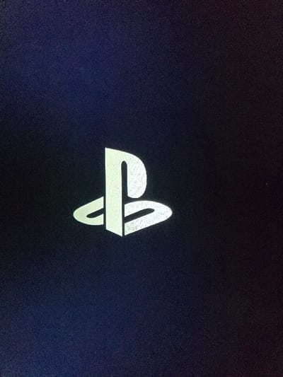 The image displays a digital screen showing the white, stylized PlayStation logo centrally against a dark blue-black background. The logo appears illuminated, with faint horizontal lines or pixelation visible within its structure, indicating it is displayed on an electronic panel. The dark background exhibits fine digital noise or grain. No people or other objects are visible, focusing solely on the screen display. The scene depicts the power-on or boot sequence of a PlayStation console, originating from the city of Kumanovo, North Macedonia.