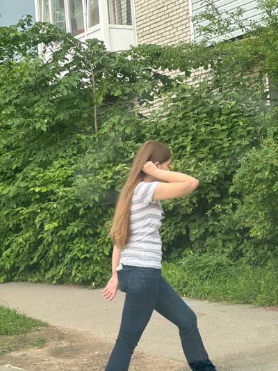 The media file shows a person from the side, standing on a paved path next to a dense wall of green foliage. The person has long, light brown hair that cascades down their back, and is wearing a striped t-shirt and dark jeans. Their right arm is bent, with their hand resting near their head, obscuring part of their face. Their left arm is extended downwards, with their hand open. The scene appears to be outdoors, with lush green bushes and grass on one side of the path, and a building with light-colored brick and large windows on the other side. The lighting suggests it is daytime. The location context is Kuzmolovskiy, Russia. There is no visible text in the image.