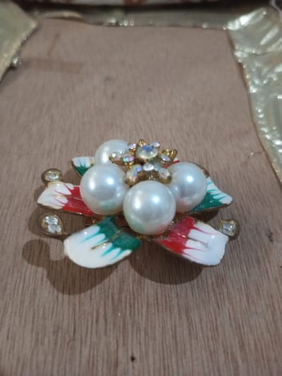 A floral brooch is centrally positioned on a light brown wooden surface with visible grain, within the city of Chandragiri, Nepal. The brooch consists of five spherical, white artificial pearls clustered together. At the center of the pearl cluster, a gold-toned metal setting holds multiple small, clear, iridescent gemstones arranged in a star or snowflake pattern. Surrounding the pearls are multiple stylized petals or leaves; some are white with red tips, while others are white with green tips. Two additional small, clear, iridescent gemstones are affixed to gold-toned bases on the outer edges of the brooch. The brooch casts a soft shadow to its bottom left. The background is out of focus, revealing a blurred light-colored, possibly gold-toned, fabric or ribbon element.