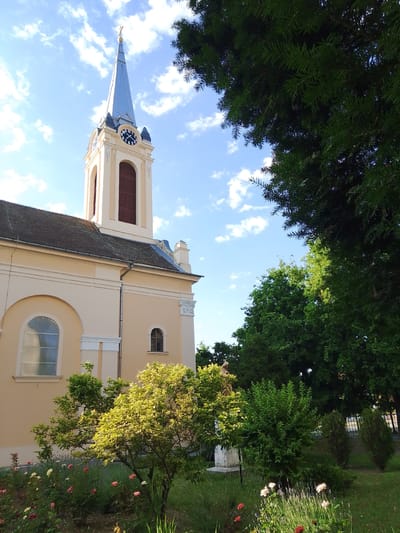 The image shows the exterior of a pale yellow church with a tall, silver-spired bell tower. The clock on the tower shows the time as approximately 10:50. The church has a dark, tiled roof and arched windows. In the foreground, there is a garden with roses in bloom, as well as various trees and bushes. The sky is blue with scattered white clouds. The scene appears to be set in a park or a quiet area of a town. Based on its architectural style and context, this church is likely located in Becej Municipality, Serbia.