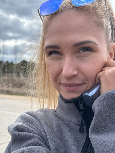 A person with light skin and blonde hair is holding a phone to their ear and smiling. They are wearing a gray fleece jacket with a black zipper. Blue sunglasses are visible on their head. The background shows a street and trees under a cloudy sky. The city of Гремяча, Russia, is not discernible from the image.