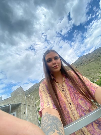 This is a low-angle selfie taken outdoors with a cloudy sky above. A woman with long brown hair, wearing a patterned button-up shirt, is the central subject. Her right arm is extended towards the camera, and a large tattoo is visible on her left forearm. In the background, a gray, unfinished brick building with a metal roof structure is situated on the left. Further in the background, a steep, barren mountain slopes upwards, partially covered in greenery. The location is identified as Chalda, Russia.